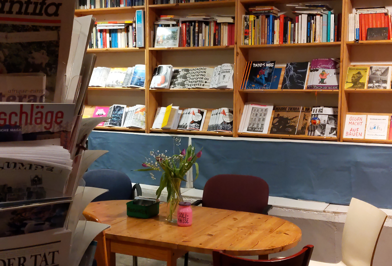 Foto vom Infoladen. Im Fokus ist ein Tisch mit Blumen. Dahinter ist ein größeres Regal mit Zines und Büchern. Im linken Vordergrund steht ein Zeitschriftenspender.